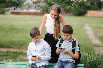 Two boys and girl use their phones during school breack. Cute boys sitting on the bench and play online games