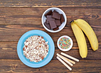 Ingredients for cooking frozen bananas in chocolate on wooden background: fresh bananas, chocolate, almond petals, sugar topping, sticks. Top view, copy space. © Natallya_ph