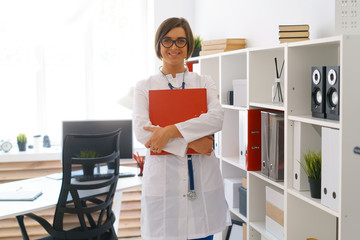 young beautiful doctor in white gown stay in her consulting room wait for patients hold binder study database of case histories
