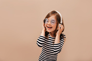 Inspired little girl in round violet glasses and stripped shirt has fun with closed eyes and broad smile 