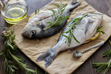 Fresh uncooked dorado or sea bream fish with lemon, herbs, oil, vegetables and spices on rustic wooden board over black backdrop, top view