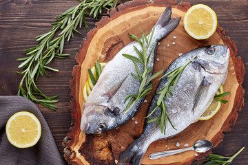 Fresh uncooked dorado or sea bream fish with lemon, herbs, oil, vegetables and spices on rustic wooden board over black backdrop, top view