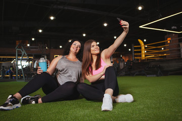 Lovely happy plus size sportswomen resting at the gym after exercising, taking selfies on smart...