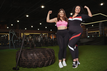 Two happy confident plus size women smiling, flexing their biceps at the gym, copy space. Happy overweight sportswomen showing their biceps to the camera. Body positive concept - Powered by Adobe