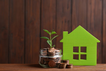 House model, jar with coins and plant on table against wooden background. Space for text