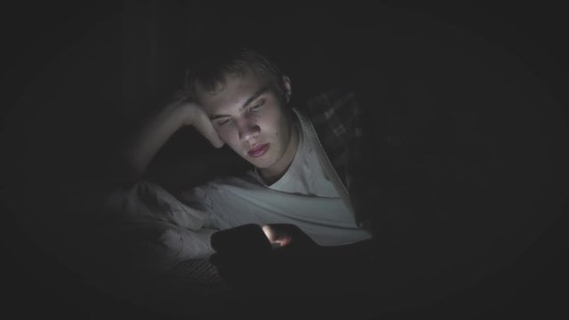 Bored teenager lying on his bed in the dark while scrolling through his phone. The light from the phone is illuminating his face.