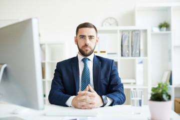 Portrait of serious confident handsome young bearded business executive sitting at table with...