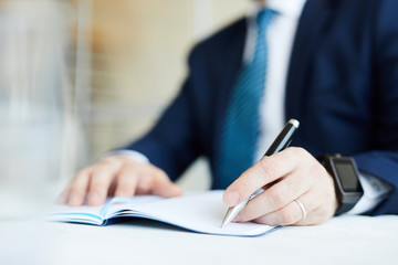 Close-up of unrecognizable businessman in suit sitting at table and making notes in diary while setting tasks for business