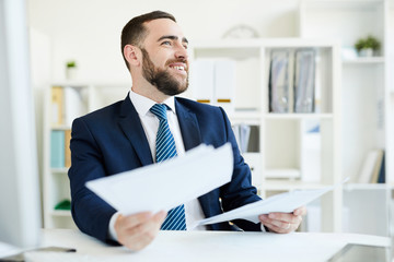 Positive excited young bearded financial manager in suit sitting at table in office and analyzing...