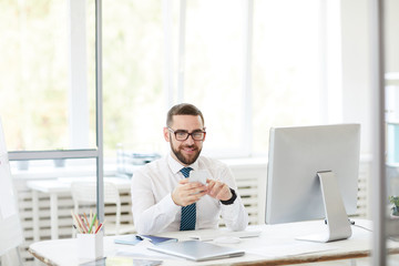 Smiling confident handsome young bearded manager in eyeglasses sitting at table and using smartphone while working in office