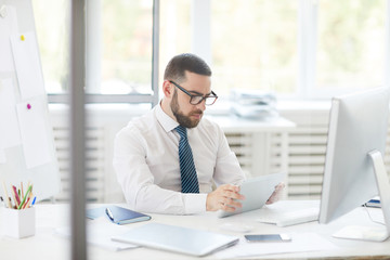 Concentrated young bearded businessman in glasses sitting at desk and analyzing online chart on tablet