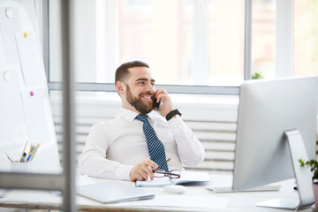 Cheerful confident young bearded manager in formal shirt sitting at table and communicating with business partner on mobile phone
