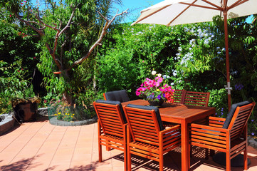 Fototapeta na wymiar Beautiful and lush summertime Mediterranean style courtyard garden with wooden table and chairs and white market umbrella.