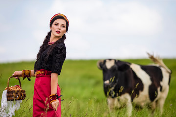 Fashion Portrait of Sensual Caucasian Brunette In Russian Kokoshnik with Basket of Bread Rings. Posing Against Cow in Field Outdoors.