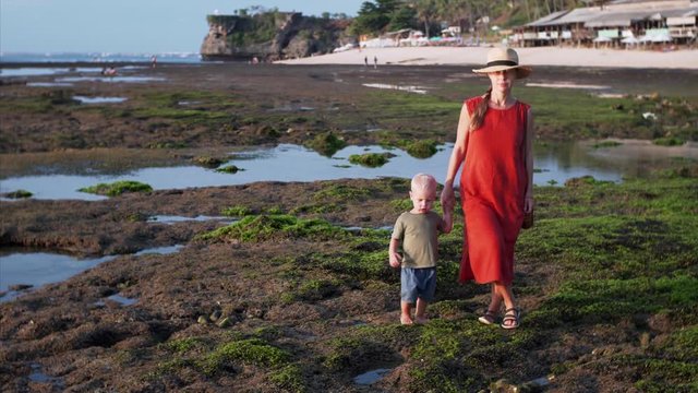 Caucasian woman in red dress is walking with barefooted son at Balangan beach at low tide. Sand is covered with green sargassa. Bali, Indonesia
