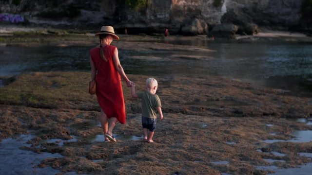 Caucasian mother and son are walking on rocky surface and puddles at Balangan beach at low tide. Bali, Indonesia