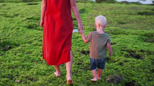 Slow motion of barefooted caucasian mother and son walking on green algae at low tide. Famous Balangan beach, Bali, Indonesia