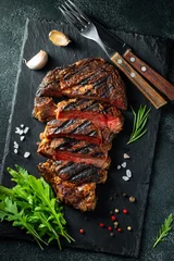 Foto auf Alu-Dibond Sliced steak ribeye, grilled with pepper, garlic, salt and thyme served on a slate cutting Board on a dark stone background. Top view. Flat lay © Vasiliy