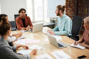 Portrait of young Middle-Eastern businessman talking to colleagues sitting at meeting table and...