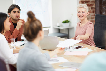 Portrait of mature businesswoman handing papers to colleagues while discussing strategy during meeting in office, copy space