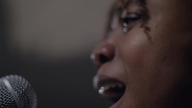 Facing a silvery and black microphone, a black African American woman young with brown skin, thick lips sings song affectionately under the bright light black singer singing in studio with emotion 