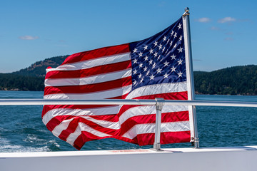 Symbol of freedom, American flag flying on the back of a boat cruising in the Salish Sea of the San Juan Islands, tree covered island and blue sky in the background