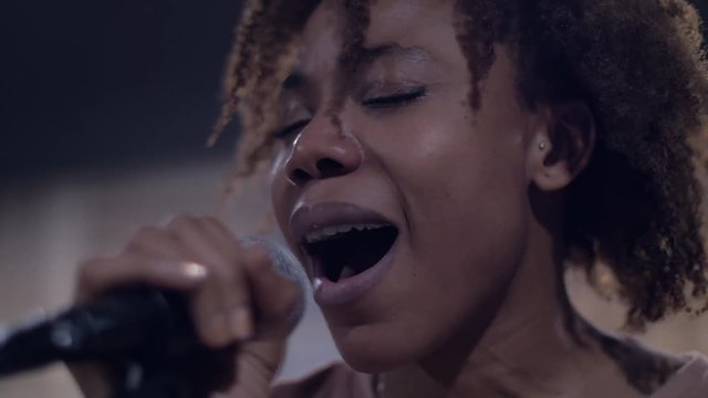 Facing a silvery and black microphone, an African American woman with brown skin, thick lips, white teeth, a flat nose and curly hair sings Rihanna's Diamonds affecitonately under the lights . 