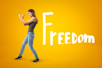 Young brunette girl wearing casual jeans and t-shirt showing double fist gesture with FREEDOM sign on yellow background