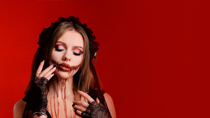 All Saints Day. Beautiful girl with terrible makeup for Halloween holds fingers of her hands near the bloody mouth on a red background. Copy space. Blank for poster or card design.