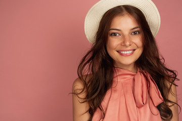 Young dark-haired woman with straw hat smiles broadly at the camera, pink background