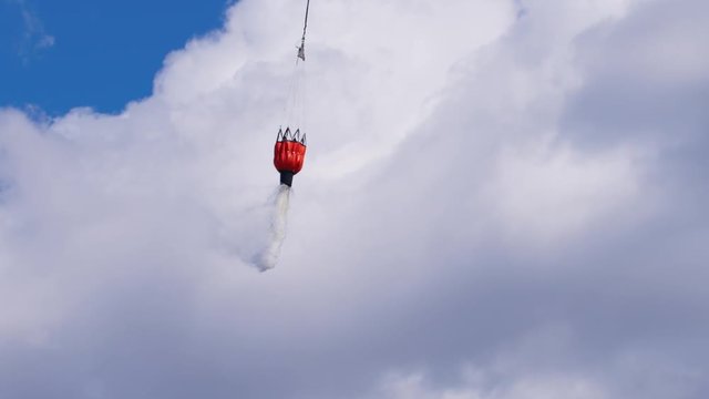 A helicopter dumps a bucket of water on a forest fire in slo-mo against a cloud filled sky.  Camera tilts down to track falling water.