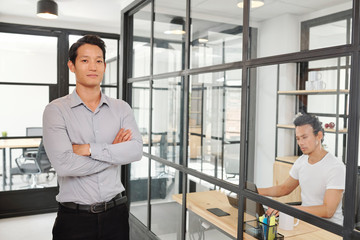 Portrait of confident young Asian entrepreneur standing in modern office with glass walls and smiling at camera