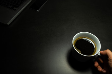 hand holding a white cup of hot black coffee against dark background with laptop and mobile phone
