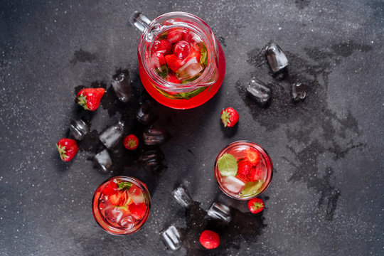 Glasses And Carafe With Strawberry Soda Drink. Cool Refreshing Homemade Lemonade Mixed Ripe Berries And Herbal Mint. Splash Of Water And Ice Around Of Cup Isolated On Black Background. Top View