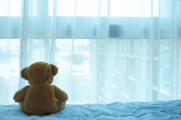 brown bear doll sitting on the bed and looking thru the curtain and window