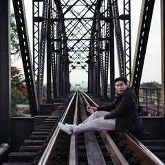 Asian tourist man in overcoat drinking takeaway coffee cup and sitting at black iron bridge railway background.