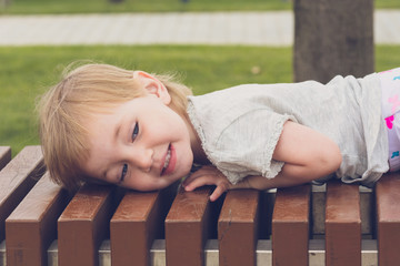 Contented child in bright clothes is lying and resting on long bench in city park.