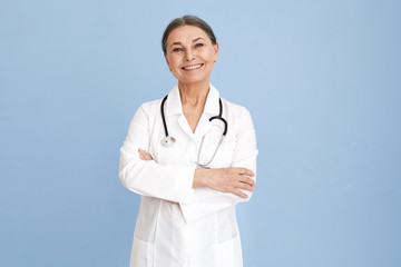 Horizontal image of happy successful mature gray haired female pediatrician posing at blue wall, keeping arms on her chest. People, age, healthcare jobs, occupations and medical careers concept