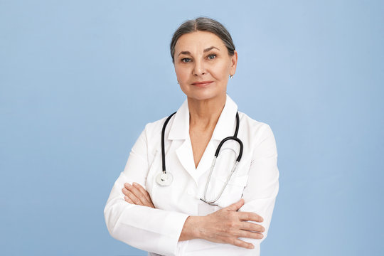 Attractive Confident Sixty Year Old Female Surgeon Wearing White Uniform And Stethoscope Keeping Arms Folded On Her Chest Posing Isolated Against Blue Wall Background With Copyspace For Your Content