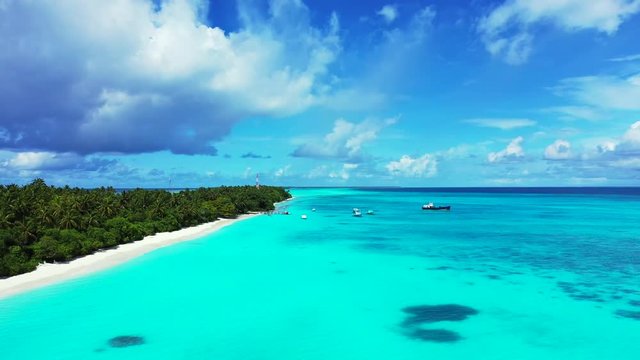 Aerial view of a flight along a tropical island. In the turquoise sea under a blue sky with some clouds. Boats in the distance. dolly in with a slight tilt up.