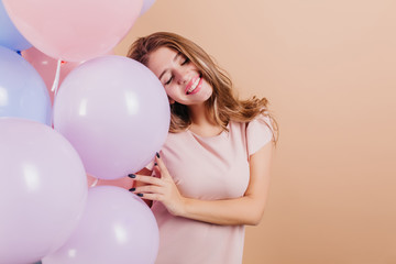 Adorable long-haired girl standing with eyes closed and holding party balloons. Studio shot of dreamy caucasian female model in pink t-shirt enjoying her birthday.