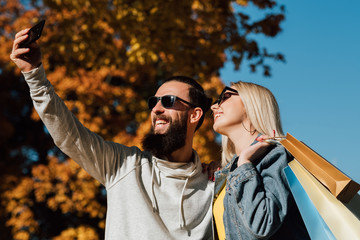 Autumn shopping travel. Couple taking selfie on smartphone. Blur fall trees and blue sky background.