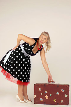 Beautiful blond woman in pinup style, dressed in a polka-dot dress, stands and lifts a travel suitcase, white background