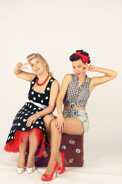 Two pinup girl friends sitting on a road suitcase on white background