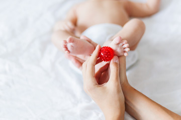 Mother holds newborn baby's feet. Tiny fingers and red massage ball in woman's hand. Cozy morning at home.