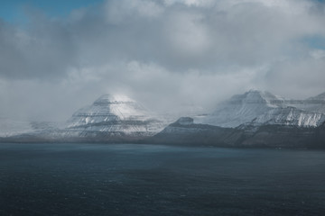 Aerial helicopter shot of snow-covered mountains at the shore of the Faroe Islands with low-hanging clouds during cold spring morning (Faroe Islands, Denmark, Europe)