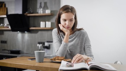 Young woman reading magazine near cup at table