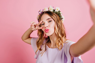 Close-up portrait of enchanting white girl wears trendy flower wreath. Charming european lady with blonde hair making selfie on pink background.