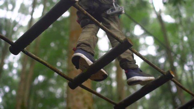 Superslowmotion shot of a little boy in a safety harness climbs on a route in treetops in a forest adventure park. He climbs on high rope trail. Outdoor amusement center with climbing activities