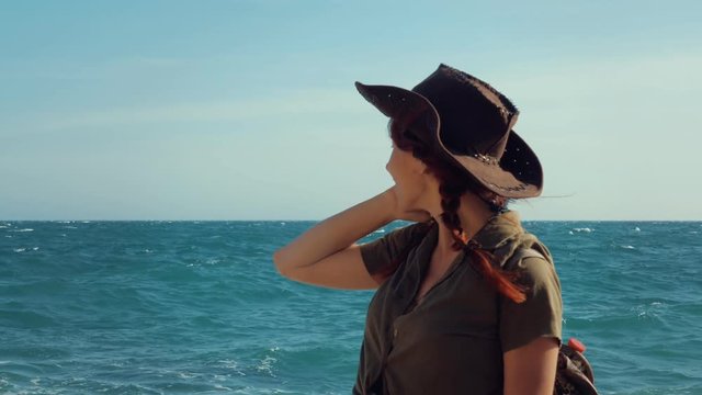 Portrait of a beautiful smiling red-haired traveler girl in a cowboy hat on the sea beach. Slow motion.